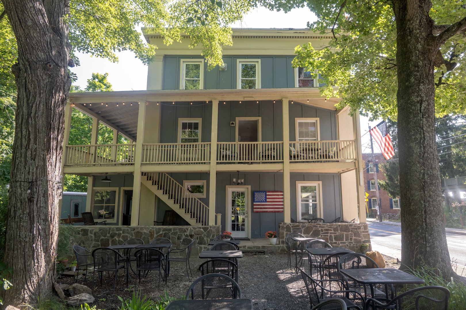 A charming three-story home nestled among mature trees, featuring a welcoming two-tier wraparound porch perfect for morning coffee or evening relaxation, with outdoor dining space shaded by leafy branches.