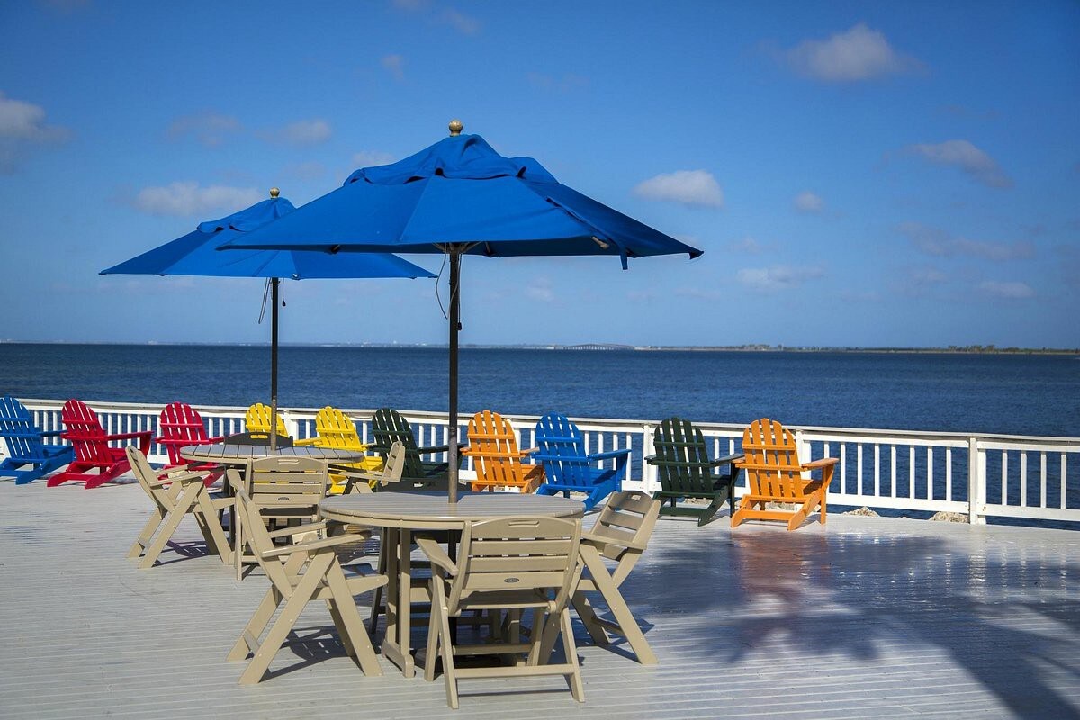 Vibrant waterside deck offers a colorful collection of Adirondack chairs and shaded dining under blue umbrellas with sweeping bay views.