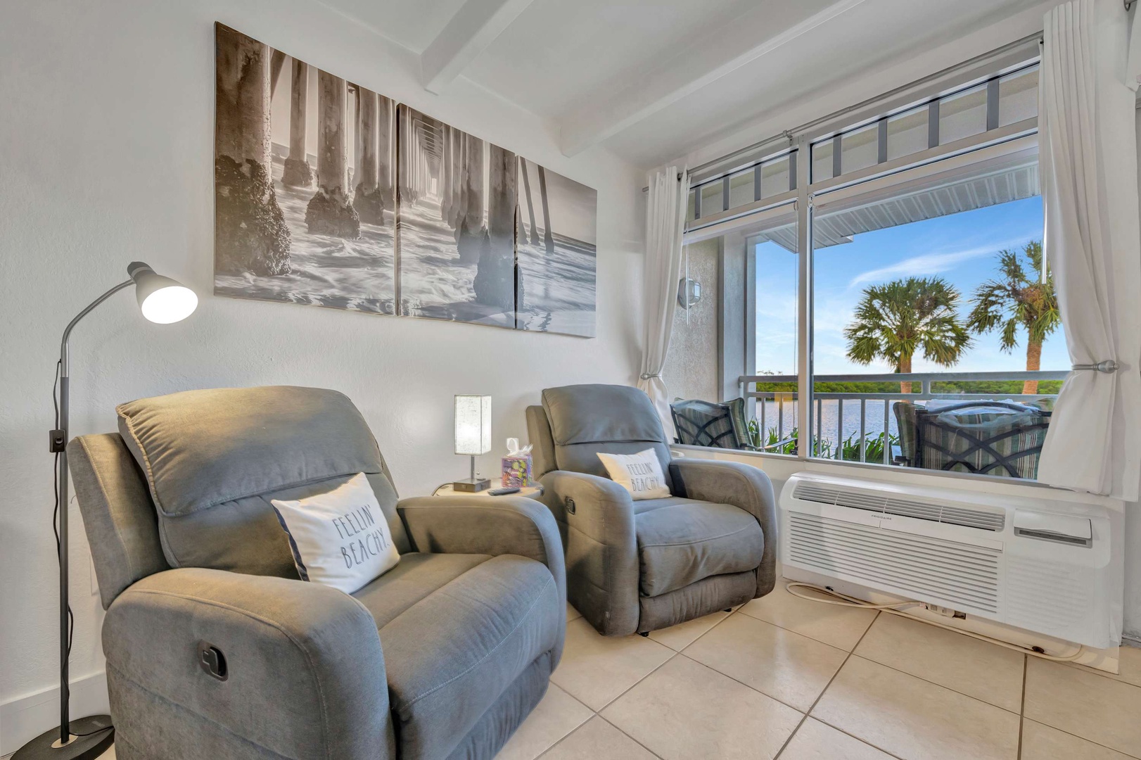 Sink into plush recliners and gaze out at the water view framed by your private balcony. Palm trees sway in the breeze as natural light fills this peaceful retreat, inviting you to unwind and soak in the coastal charm.