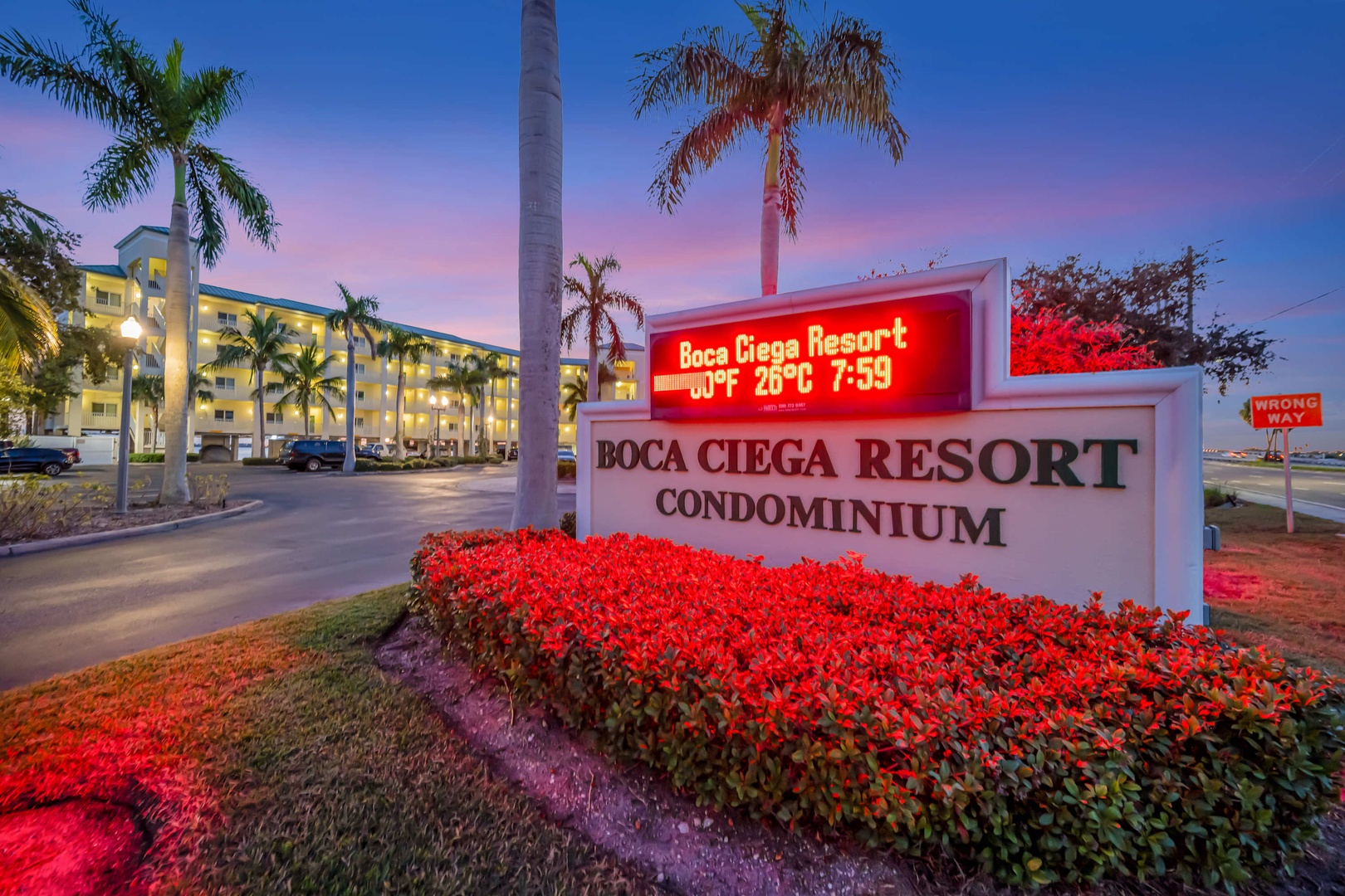 The Boca Ciega Resort Condominium entrance sign welcomes arrivals, illuminated against a tropical Florida sunset with swaying palms and the main building in the background.