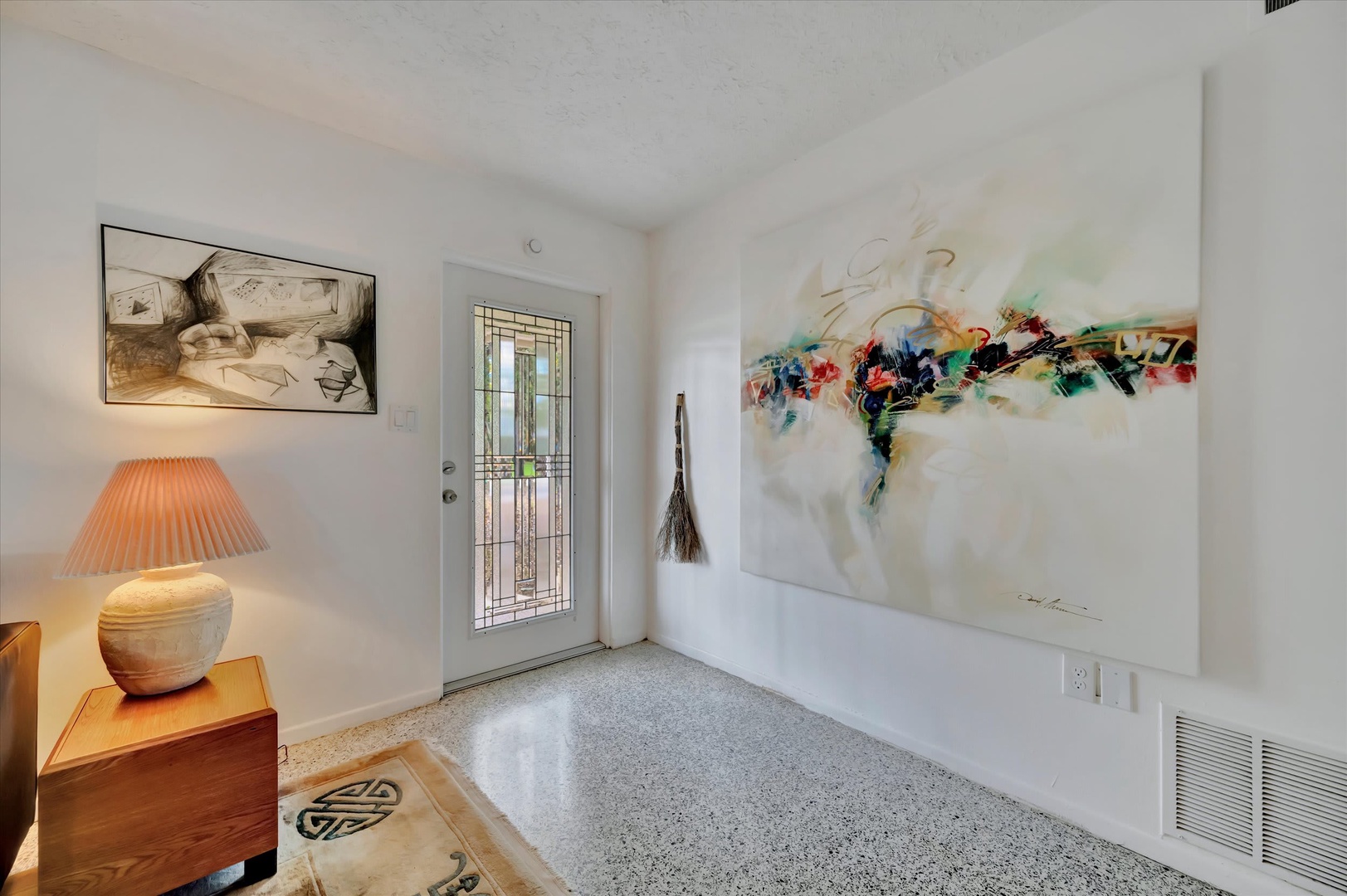 Step into a bright, art-filled entryway where natural light streams through a stained glass door, setting a welcoming tone for your stay. The curated artwork and warm lamplight create an inviting first impression.