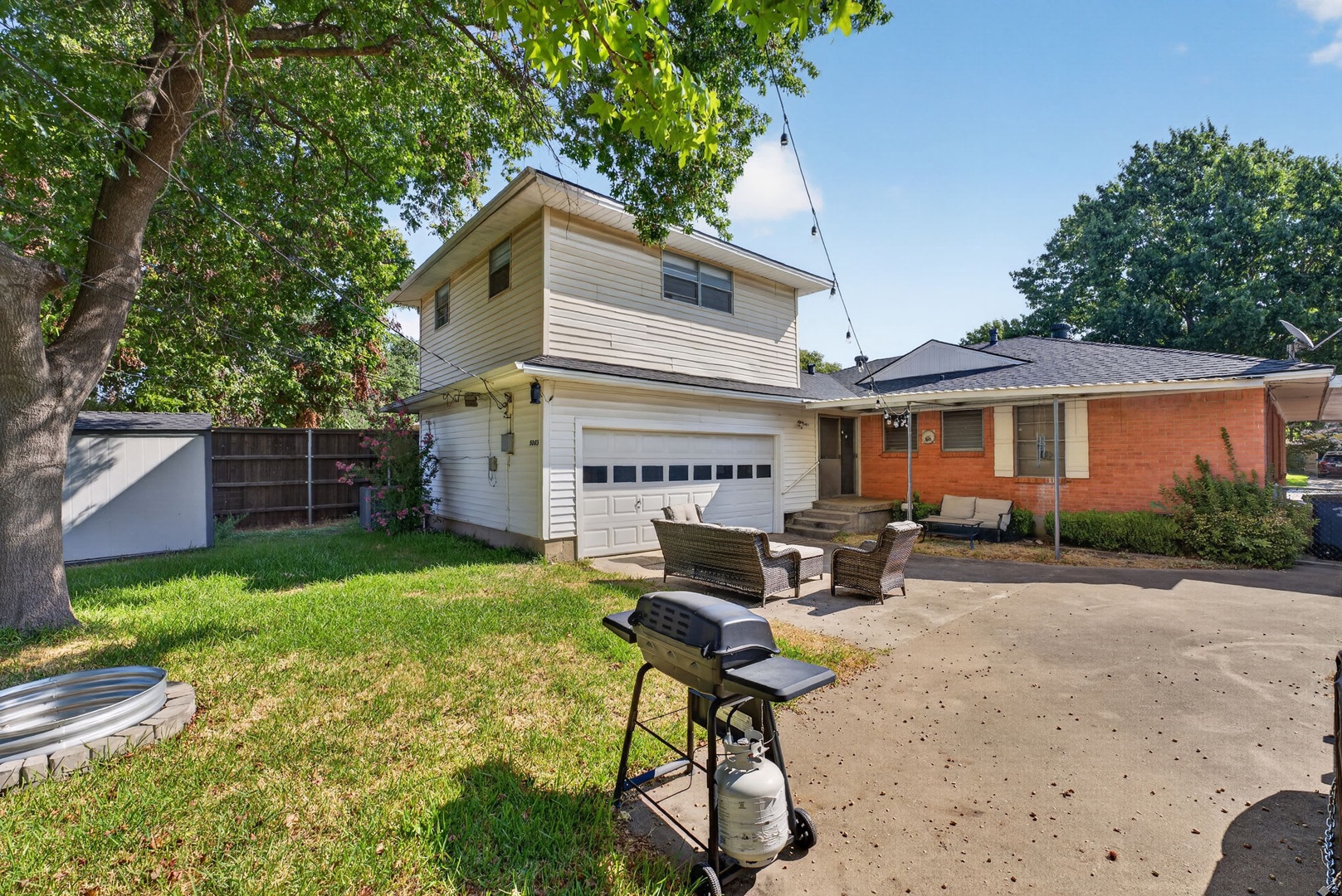 The property exterior in a quiet neighborhood offers mature shade trees, a spacious driveway, and a backyard patio with a grill for relaxed outdoor dining.