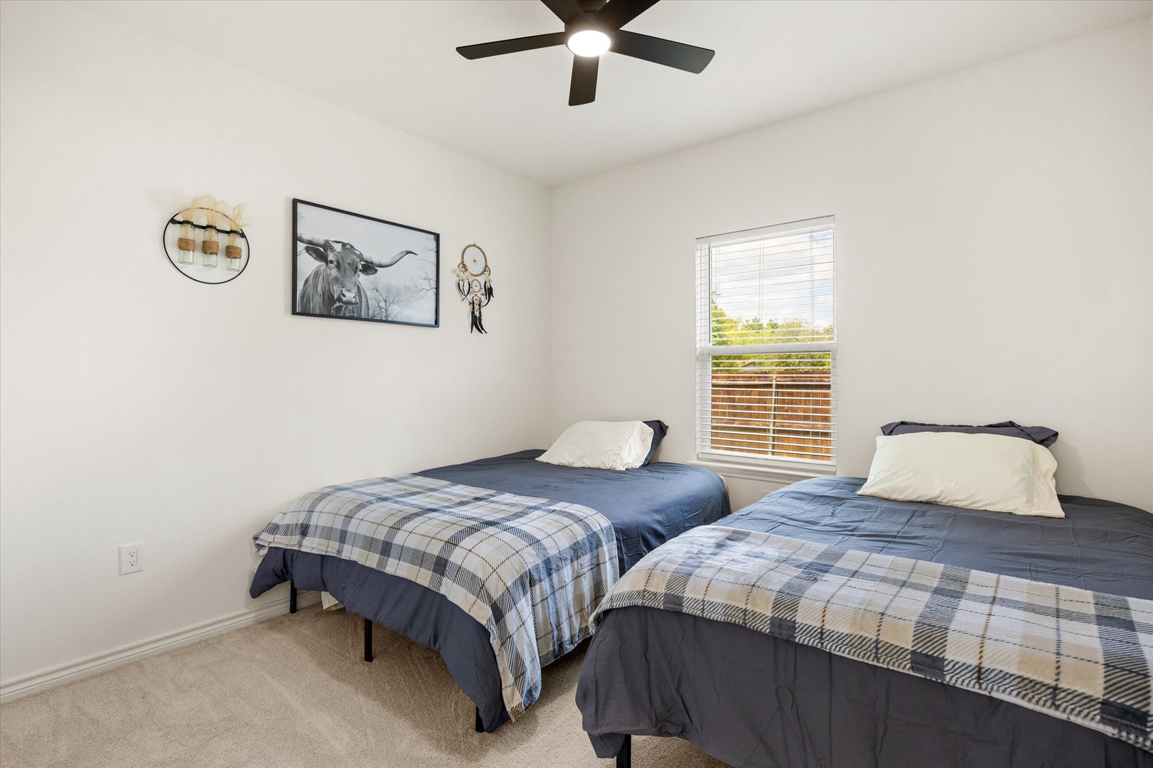 Your peaceful bedroom features two comfortable twin beds dressed in soft linens, a gentle ceiling fan to keep you cool, and natural light from the window overlooking open fields. Wake up to serene countryside views.