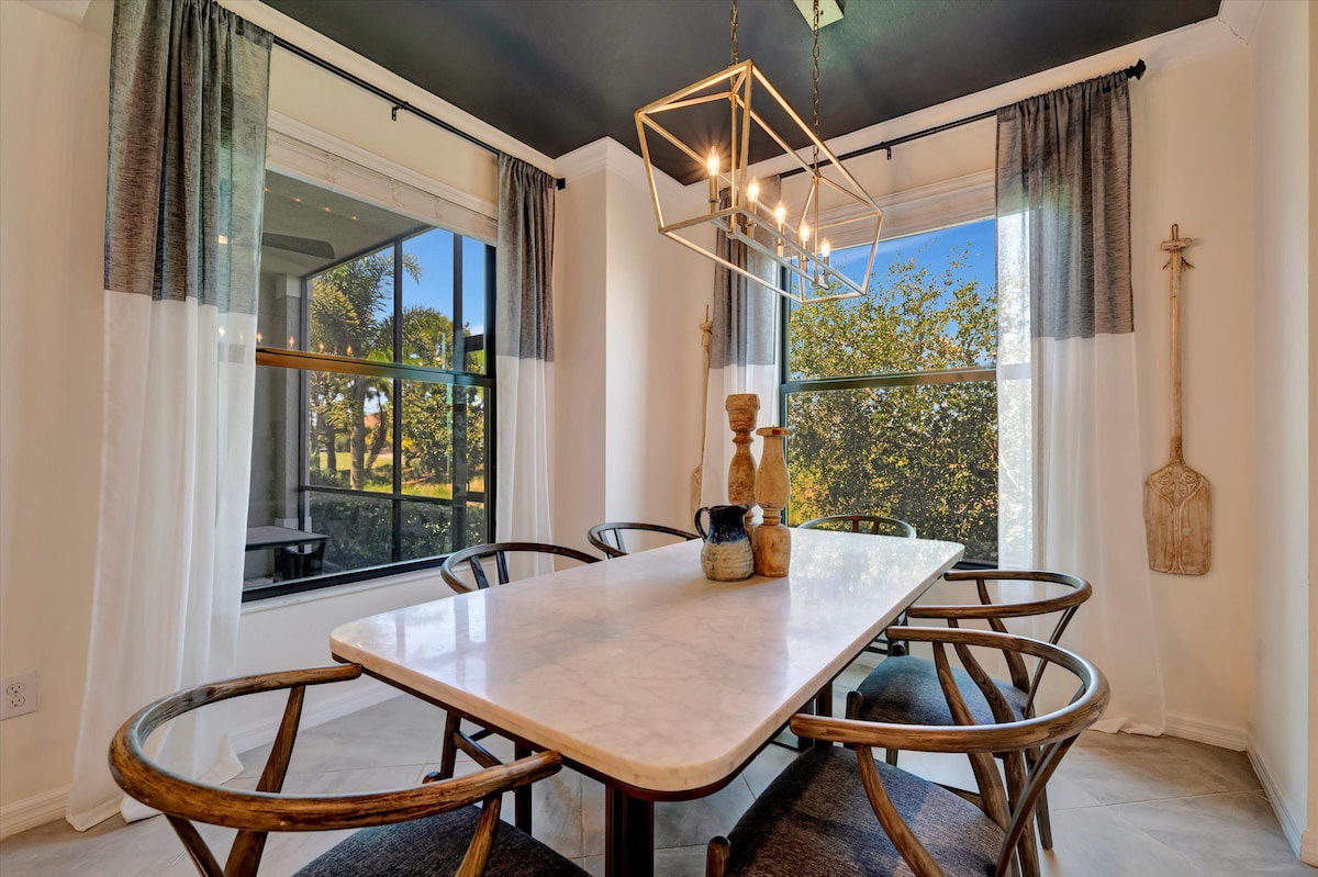 Gather around this elegant marble table under a modern chandelier with sunlight streaming through windows framed by lush greenery. Your mornings and shared meals feel special here.