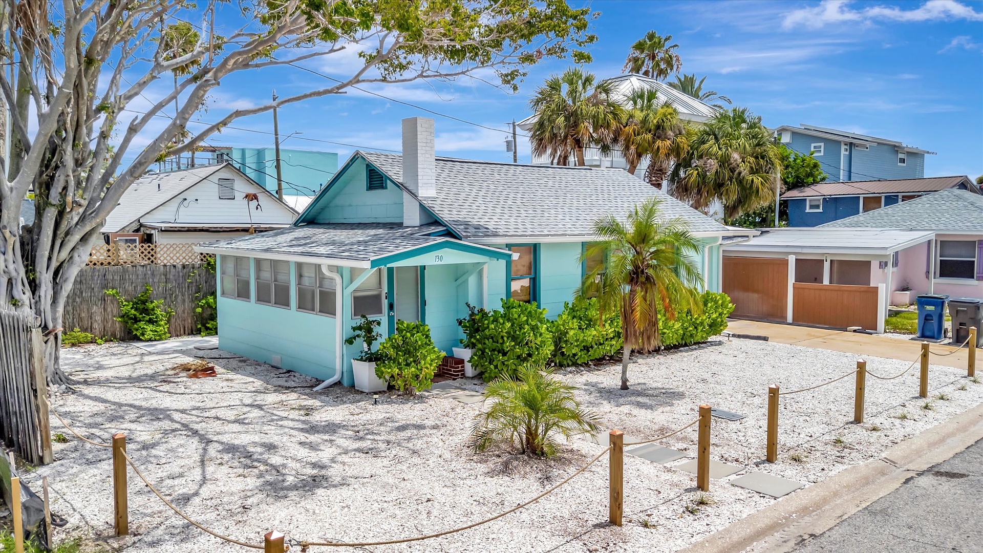 A charming coastal cottage in a residential beach neighborhood, featuring tropical landscaping and convenient parking. The property is surrounded by mature palm trees and lush greenery, offering a peaceful setting just steps from nearby amenities.