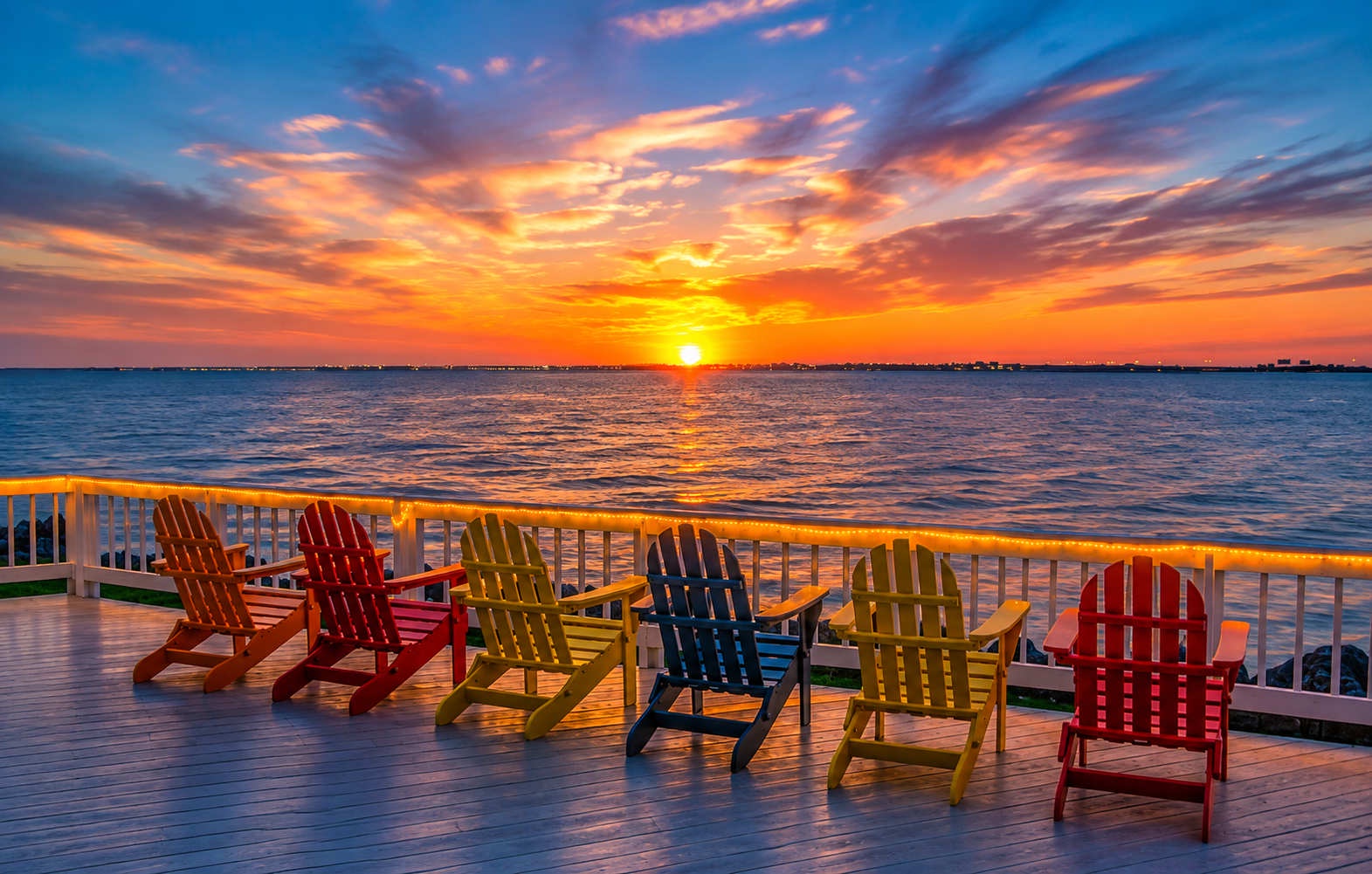 A stunning waterfront deck with colorful Adirondack chairs awaits at sunset, where brilliant golden skies meet calm waters along the property's shoreline.