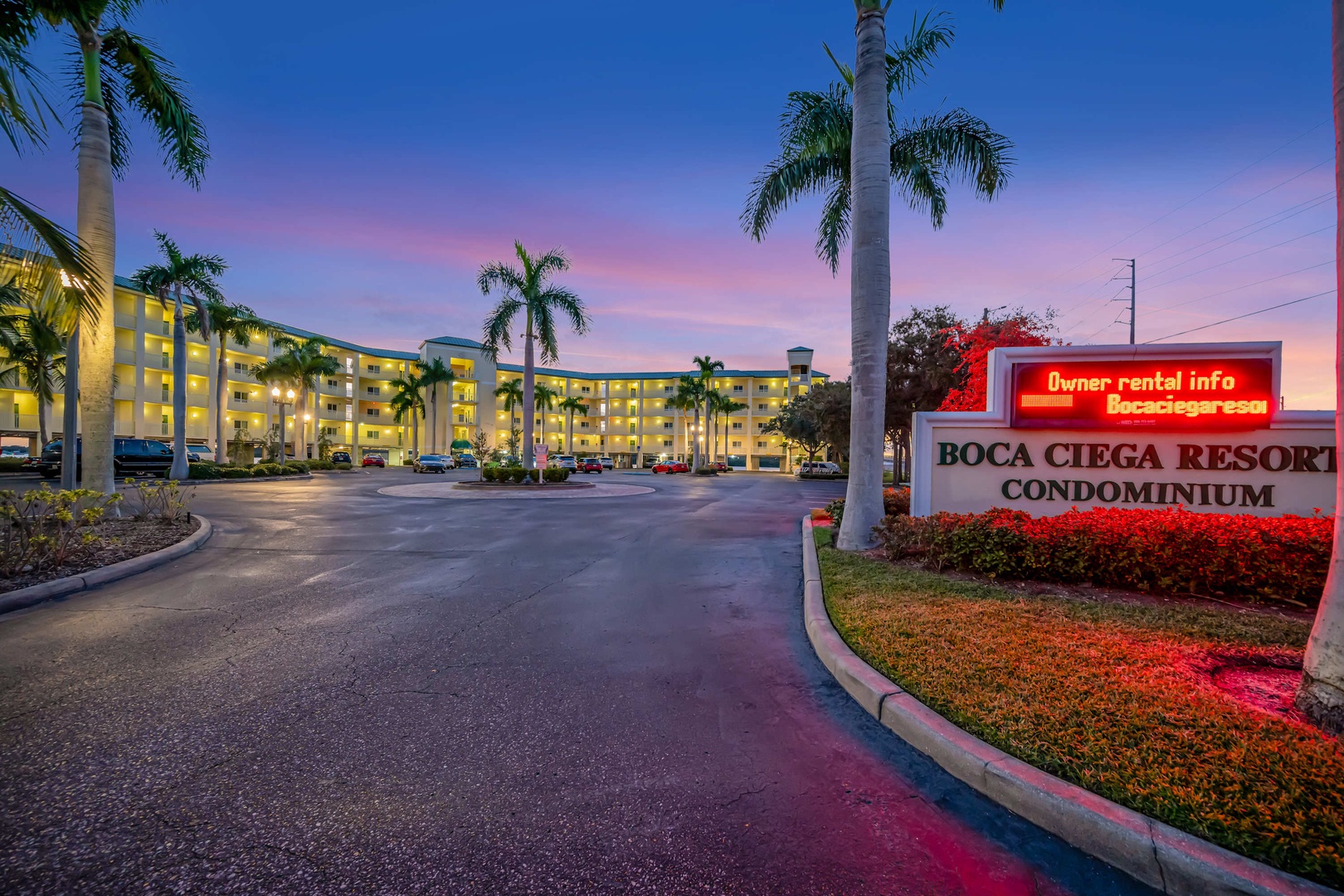Boca Ciega Resort Condominium welcomes visitors with its illuminated facade and tropical palm-lined entrance, captured during a stunning sunset hour.
