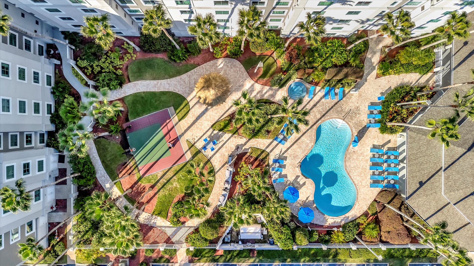 Aerial view of the property's lush tropical courtyard, featuring two sparkling pools, tennis court, and abundant palm trees surrounded by the residential buildings.