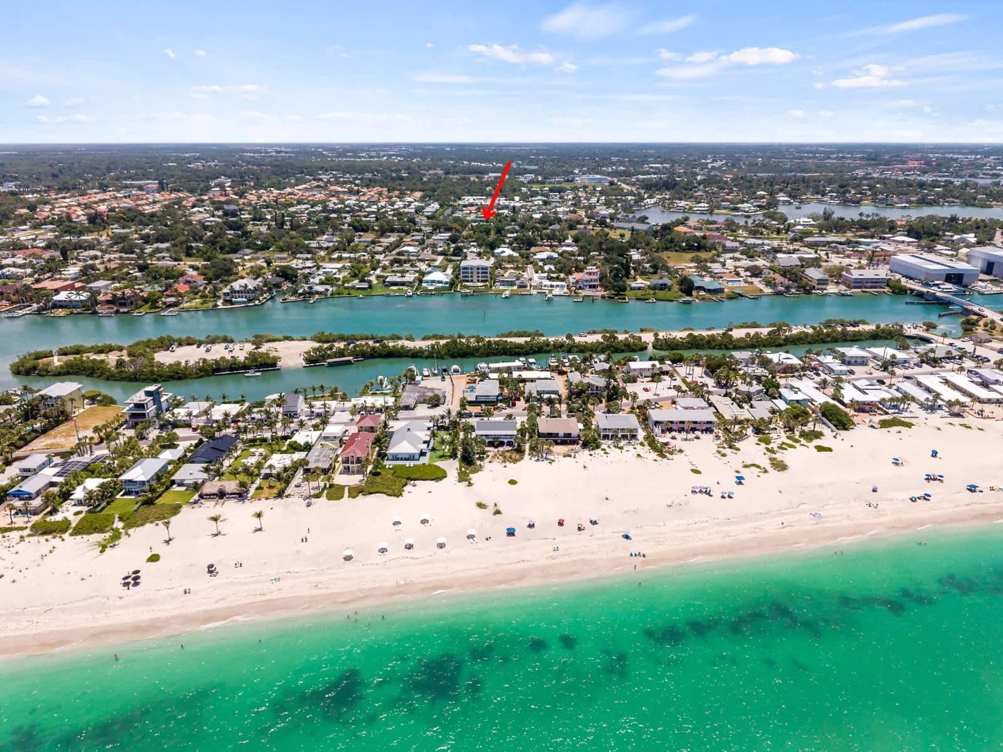 Aerial view capturing a beachfront neighborhood with sandy shores, turquoise waters, residential streets, and lagoon-style waterways extending into the surrounding community.