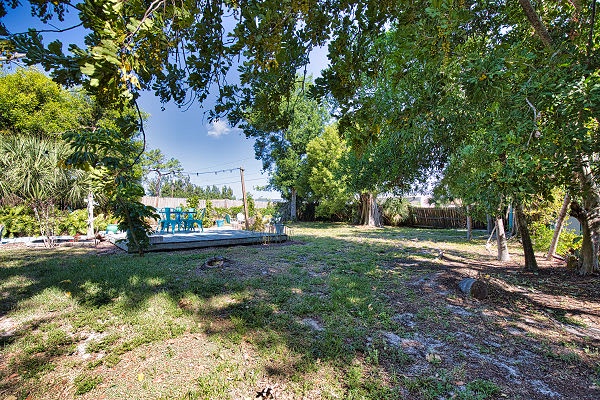 The property features a spacious backyard shaded by mature trees, with a glimpse of the waterfront and blue loungers visible in the background.