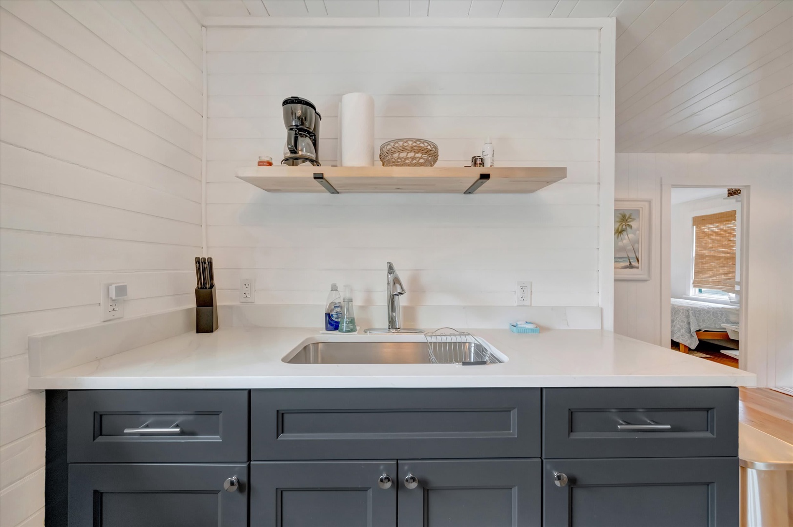 Your well-appointed kitchenette features sleek countertops, a modern sink, and ample storage. Crisp white shiplap walls and open shelving add coastal charm, while natural light invites you to prepare meals in comfort.
