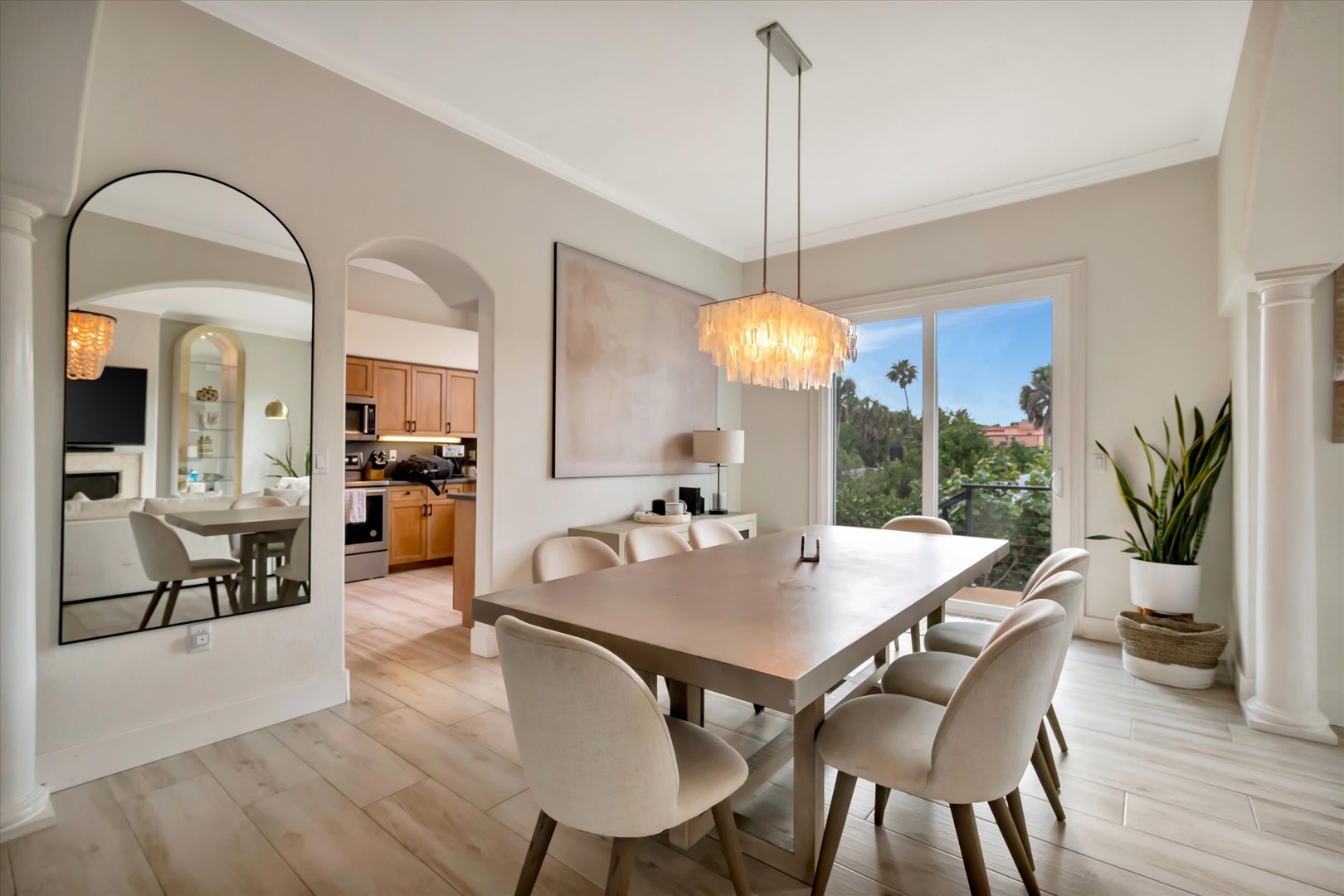 Gather around the warm wood dining table beneath a statement chandelier. Soft natural light flows through large windows framing palm trees outside, creating an inviting space where your group can share meals and memories.