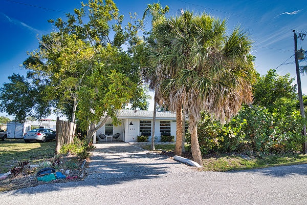 The property sits nestled among lush tropical landscaping, with a charming gravel driveway leading you to the entrance beneath sunny Florida skies.