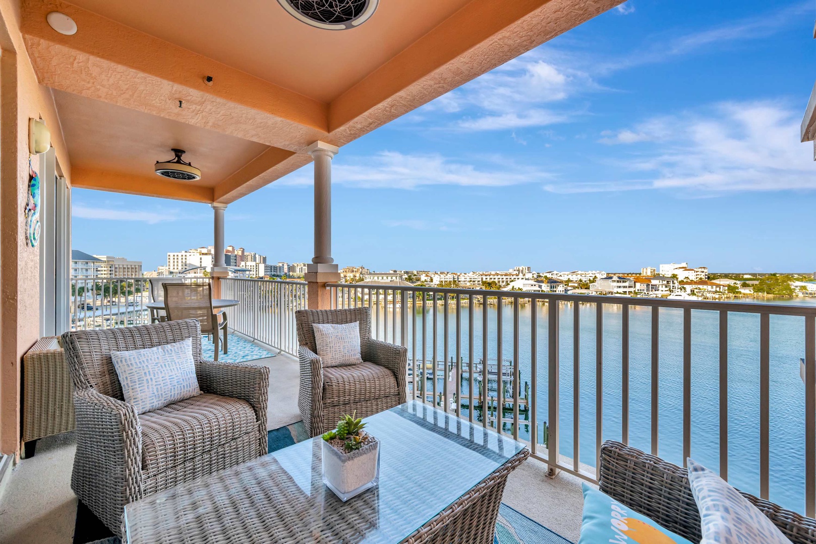 Settle into your private waterfront balcony with wicker seating and breathtaking views across the bay to coastal buildings and blue skies—perfect for morning coffee or sunset relaxation.