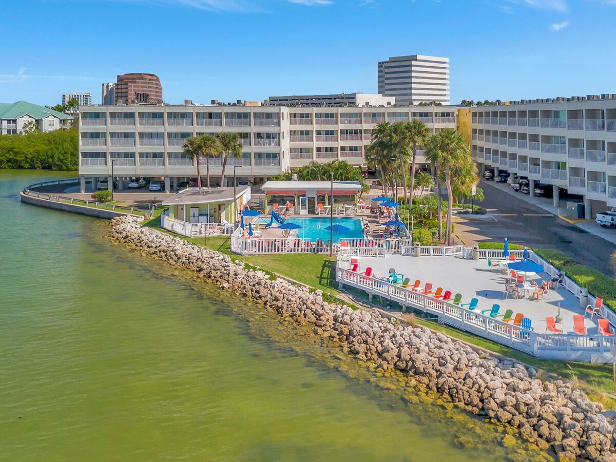 Waterfront vacation property featuring a sparkling pool, colorful lounge chairs, and palm-lined grounds against a backdrop of city skyline and calm bay waters.