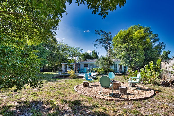 The property sits peacefully among lush greenery under bright blue skies, offering a serene retreat surrounded by mature trees and thoughtfully landscaped grounds with charming garden touches.