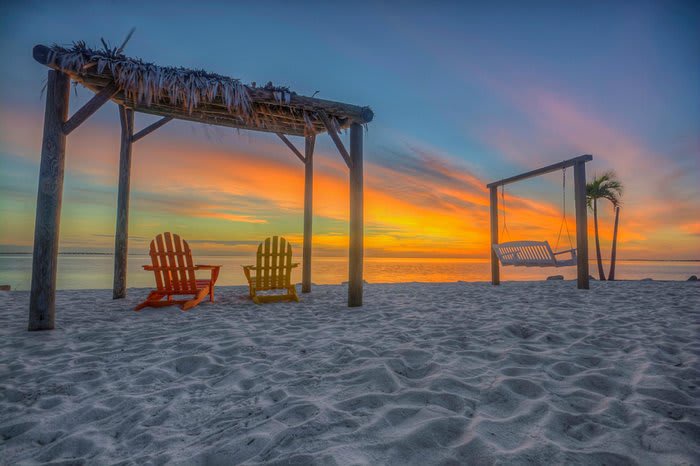 Beachfront property featuring private sandy access, shaded seating pergolas, and a coastal swing area, with stunning sunset views over the ocean.