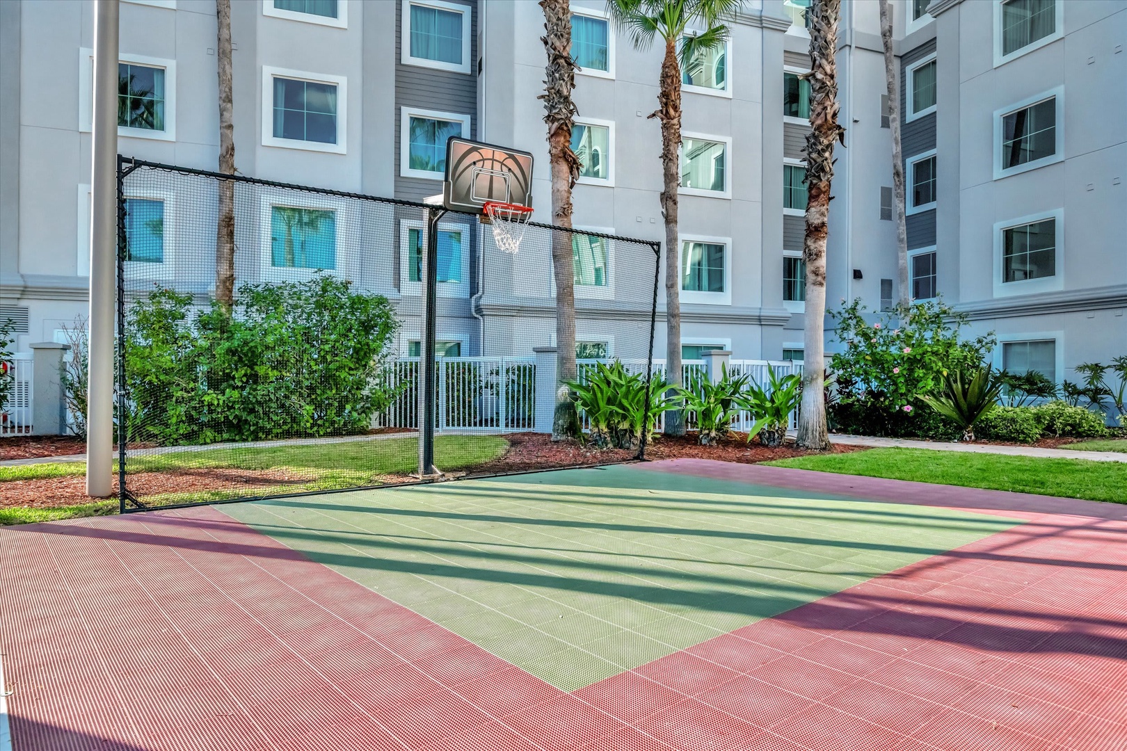 A multi-sport court set within a lush courtyard, featuring palm trees and green landscaping with the property building as a backdrop.