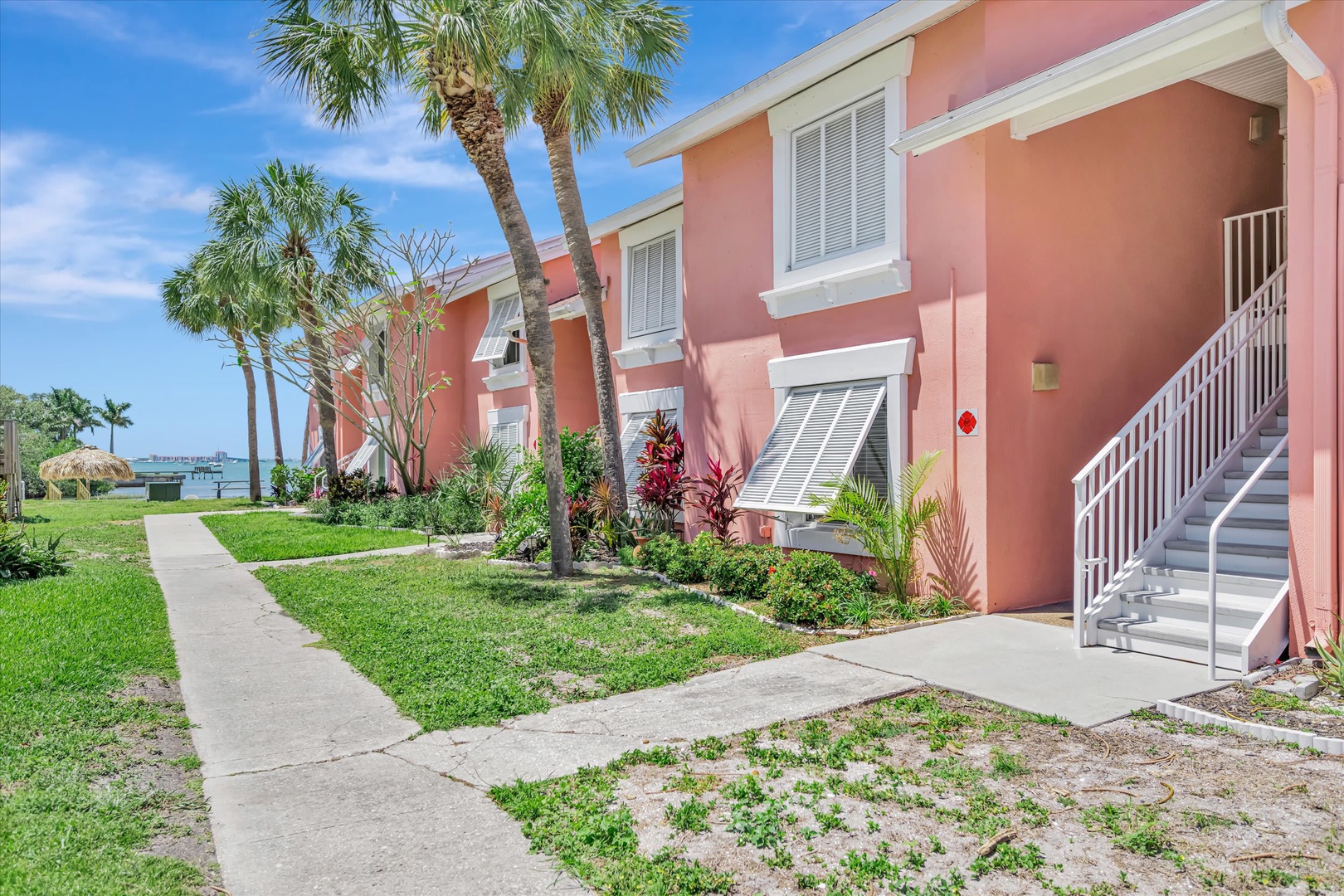 A charming coral-pink building framed by swaying palms and lush tropical landscaping, with the water visible just beyond—a welcoming seaside setting for your stay.