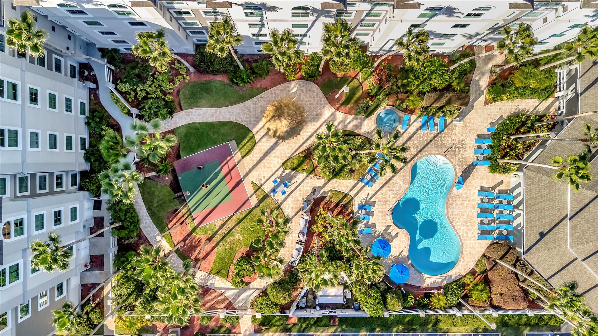 An aerial view showcases the property's tropical landscaping, featuring a sparkling pool surrounded by loungers, palm trees, and lush gardens within a multi-building complex.