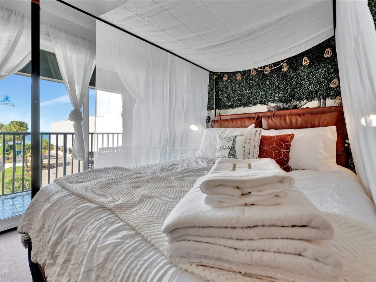 Drift off beneath soft curtains in a dreamy bedroom with plush linens and a lush green accent wall, while fresh towels and gentle light from your private balcony invite pure relaxation.