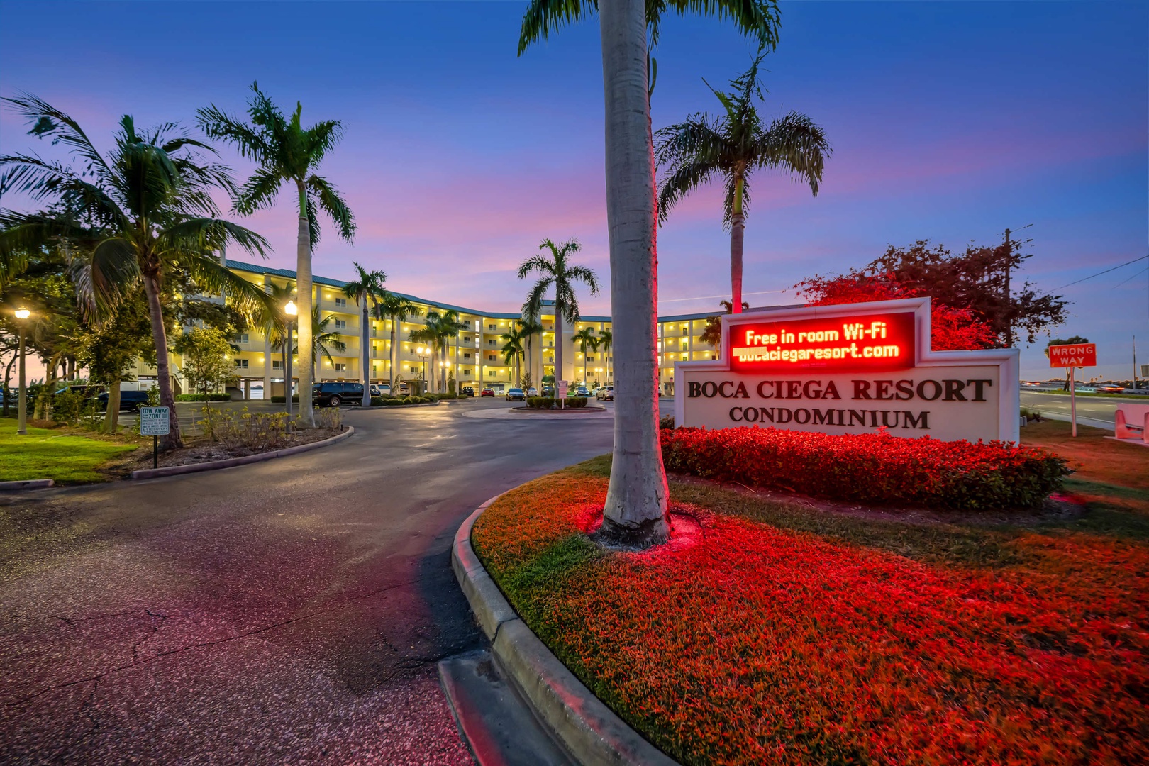 Boca Ciega Resort Condominium welcomes guests with illuminated signage and palm-lined entrance at twilight, marking the arrival to this waterfront destination.