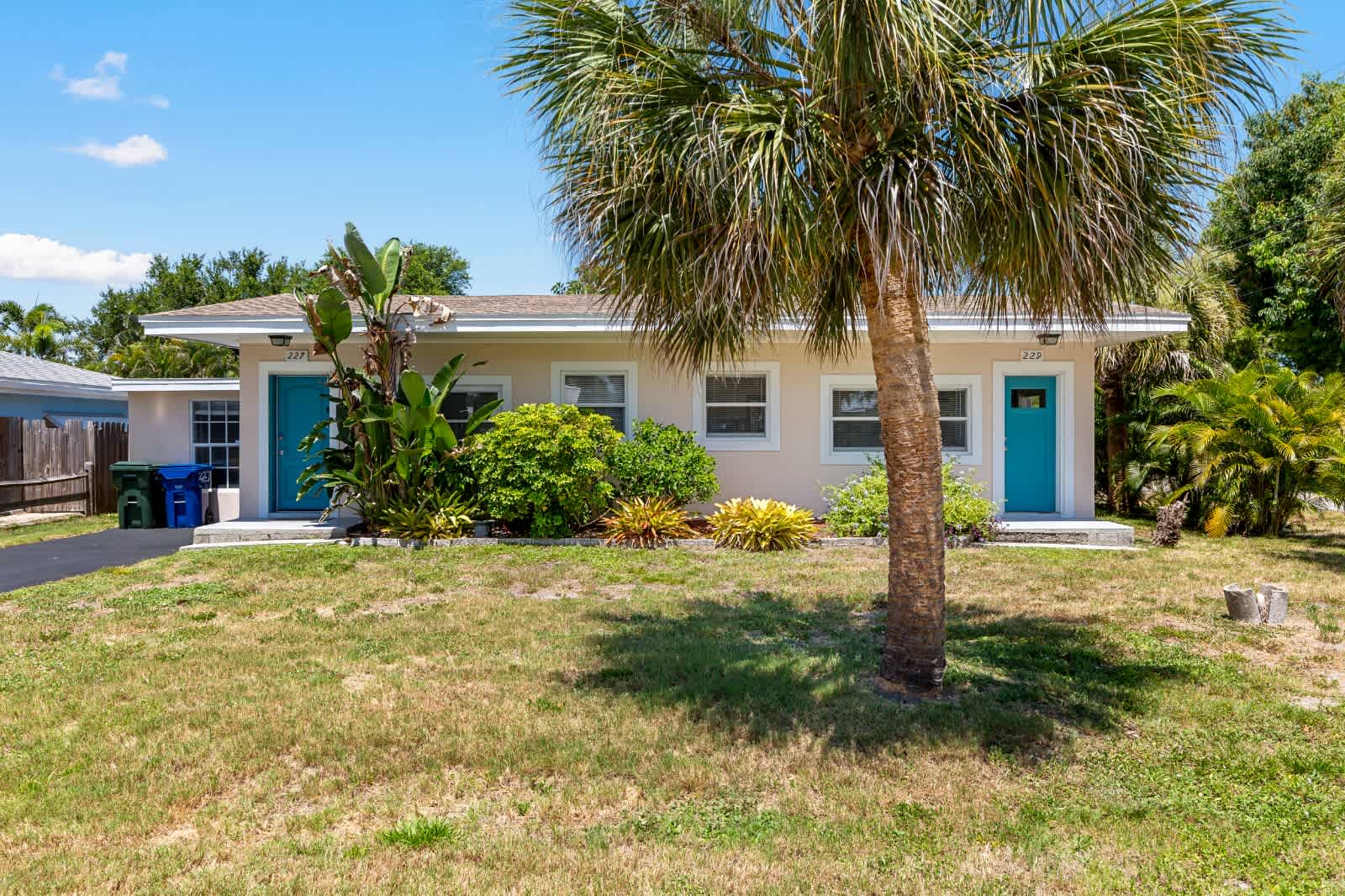 A charming single-story property welcomes you with bright turquoise doors and lush tropical landscaping, featuring a tall palm tree and vibrant greenery. This Florida-style home offers a peaceful retreat in a sunny, residential neighborhood.