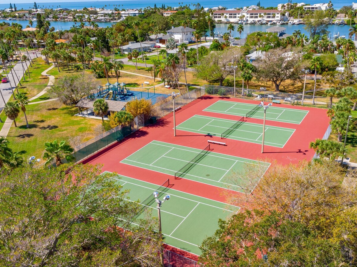 Aerial view of the property's tennis courts, playground, and surrounding waterfront neighborhood with marina access and coastal scenery.