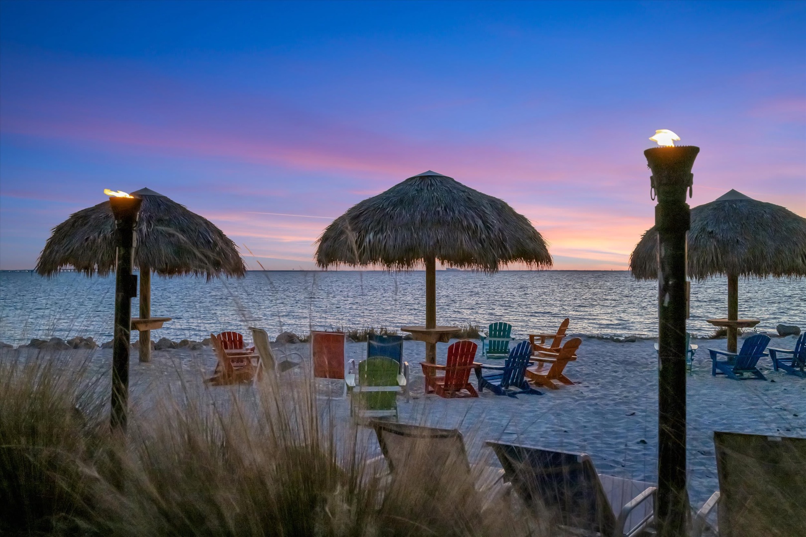 Private beachfront paradise as dusk paints the sky. Colorful Adirondack chairs and tiki torches await under thatched palapas, where your evenings unfold to the gentle rhythm of the waves.