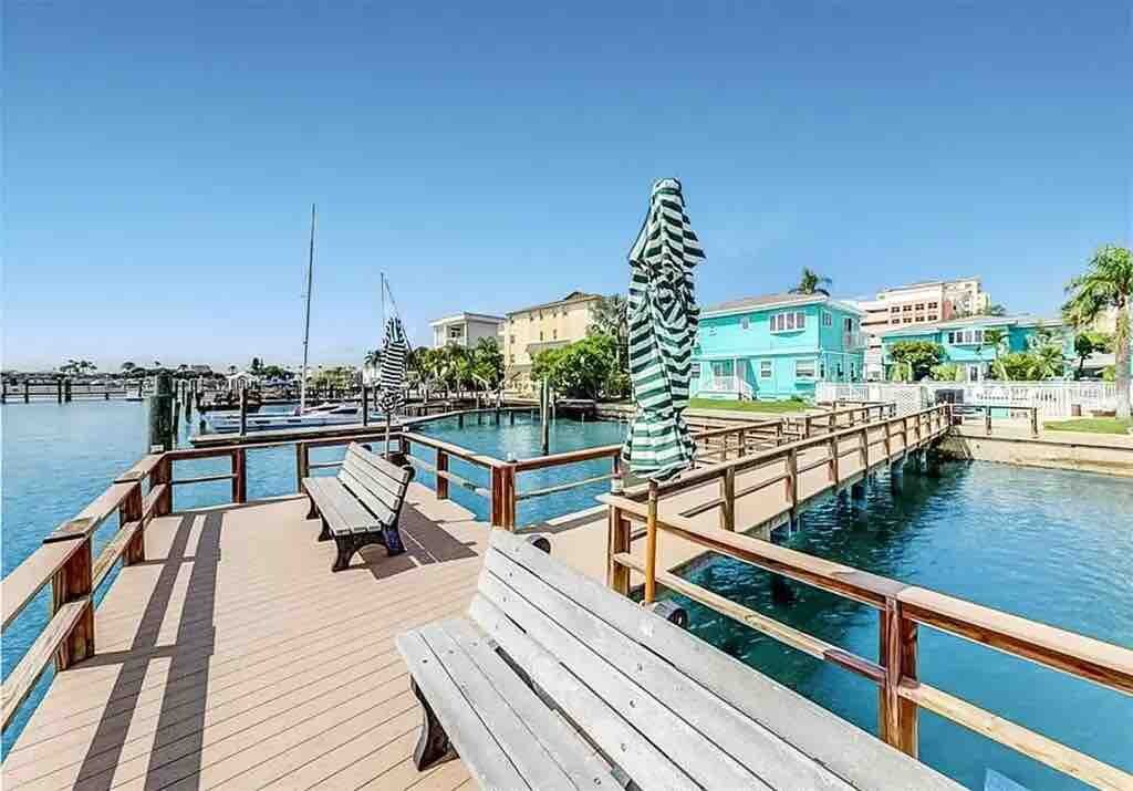 Waterfront boardwalk with boat slips and seating leads to colorful coastal buildings, offering direct water access and tropical island living.