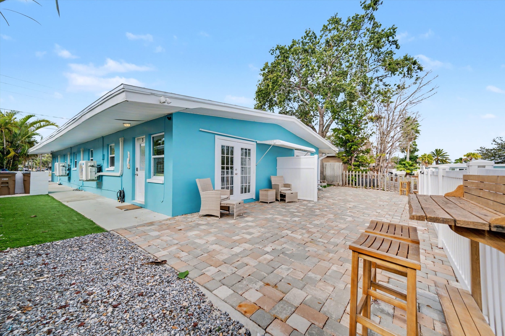 A bright turquoise beach house welcomes arrivals with charming coastal style. The spacious paver patio and lush lawn create an inviting outdoor retreat where sunny days and warm evenings await.
