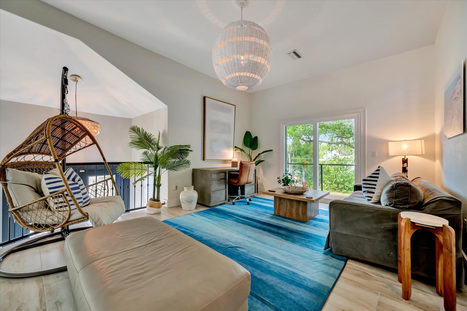 Settle into your bright, airy living space where coastal style meets modern comfort. Sink into the plush seating, sway in the woven egg chair, and let natural light stream through expansive windows overlooking lush greenery.