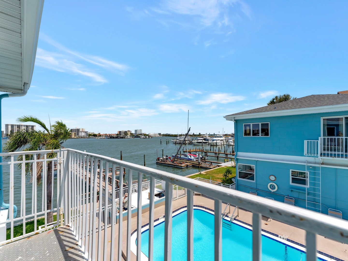 From the balcony railing, you'll see sparkling blue water, boats docked nearby, colorful beach homes, and a refreshing pool below—your perfect coastal escape.
