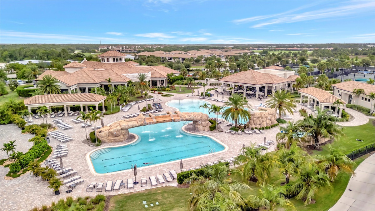 Aerial view of a resort-style property featuring a tropical oasis with multiple pools, palm trees, and Mediterranean-inspired architecture surrounded by lush landscaping and residential community.