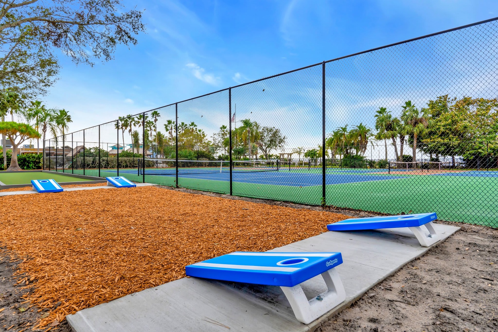 The property features tennis courts surrounded by palm trees and outdoor recreation areas. Courts are framed by a landscaped setting with natural mulch borders and distinctive blue benches.
