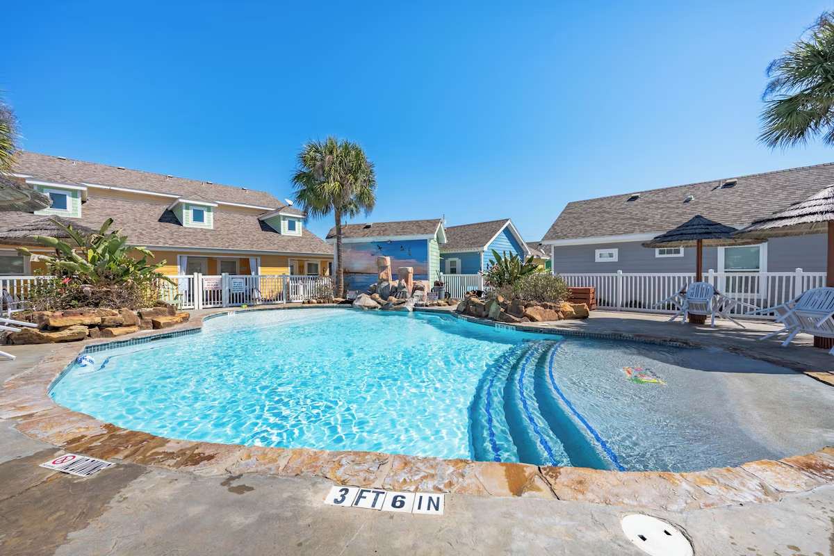 Crystalline waters invite you to unwind in this resort-style pool, surrounded by charming coastal cottages and swaying palms under endless blue skies.