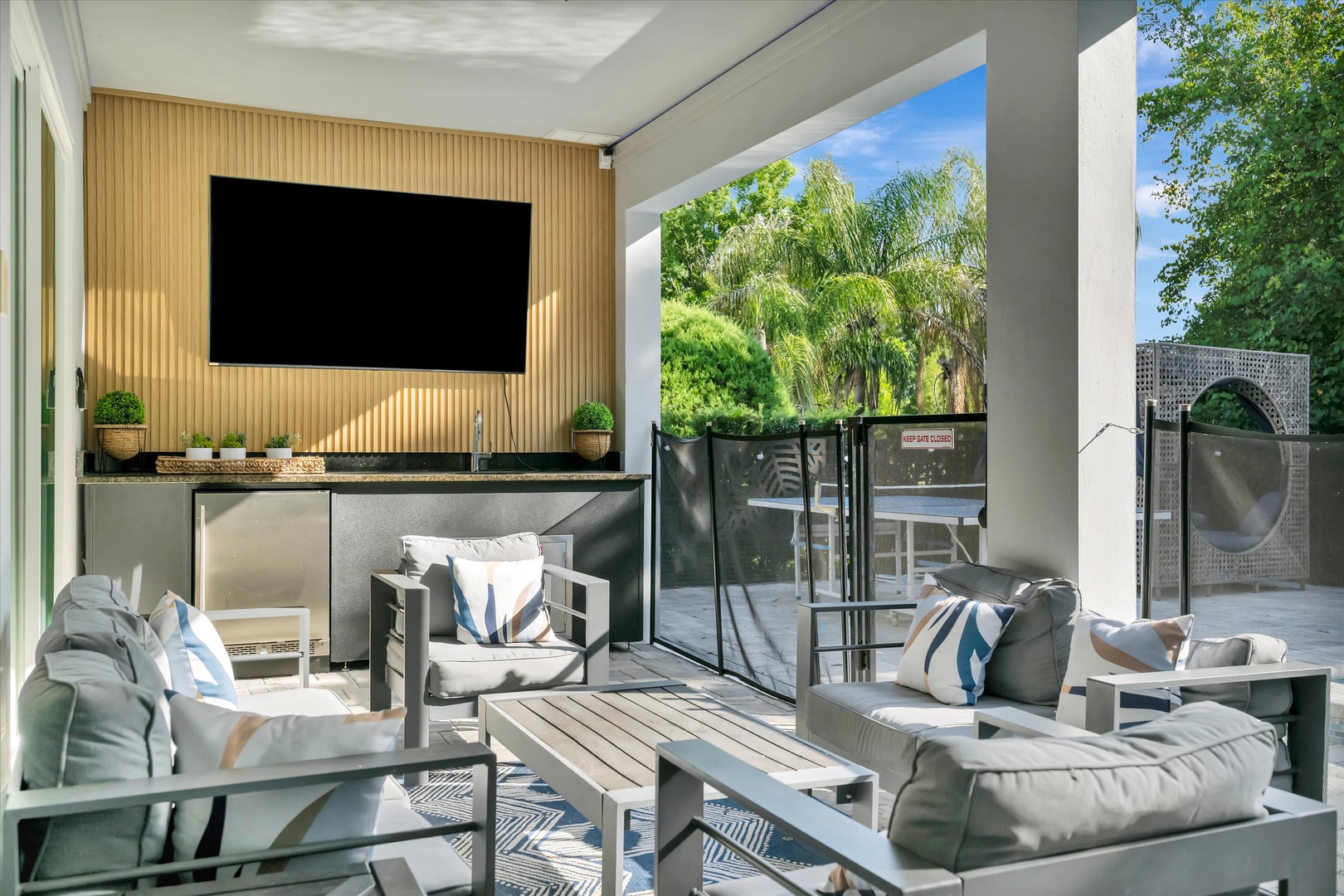 Settle into your covered patio and enjoy a seamless indoor-outdoor flow with modern furnishings, a large entertainment wall, and lush garden views just beyond the glass doors.