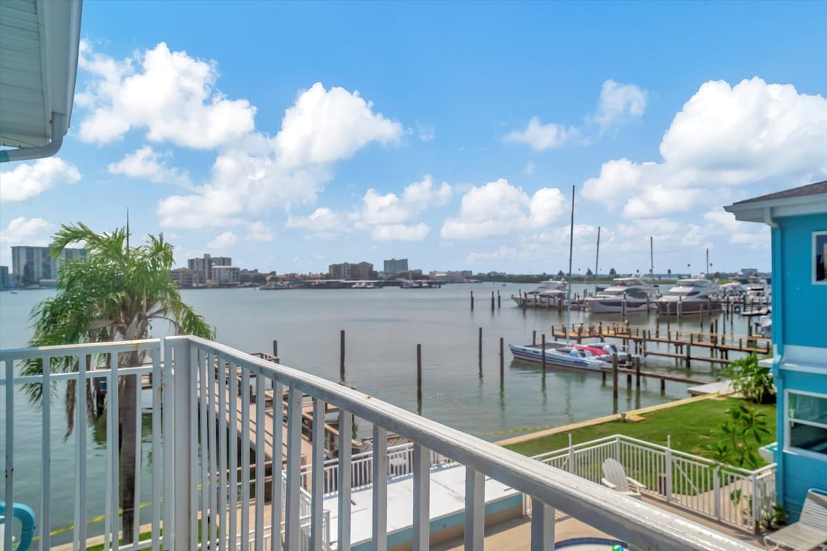 Waterfront property featuring a marina, boat docks, and distant city skyline across calm bay waters under a bright, cloud-filled sky.