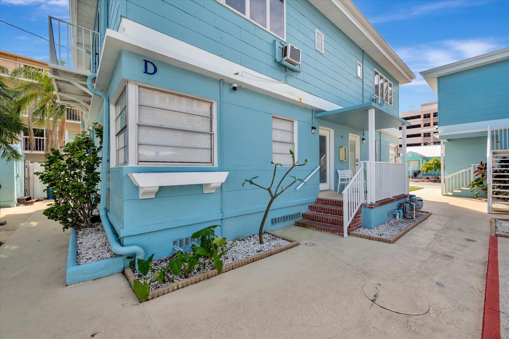 Bright coastal cottage exterior marked as Unit D, featuring white trim and a welcoming entrance surrounded by tropical landscaping in a charming beach community.
