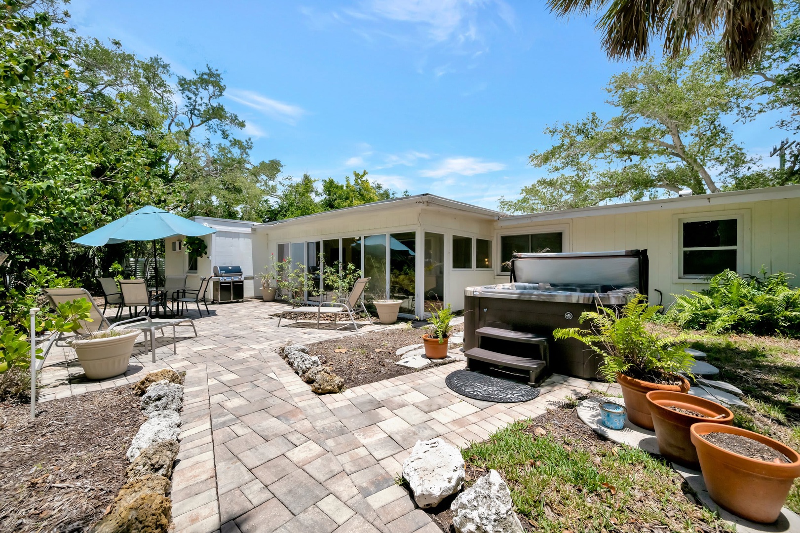 Your private backyard oasis awaits, featuring a bubbling hot tub, outdoor dining area with grill, and lush landscaping under sunny skies—perfect for relaxing or entertaining.