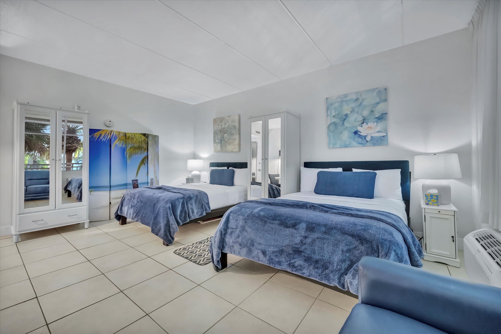 Relax in your peaceful retreat with comfortable twin beds dressed in soft blue linens, calming coastal artwork, and gentle natural light flowing through the glass doors.