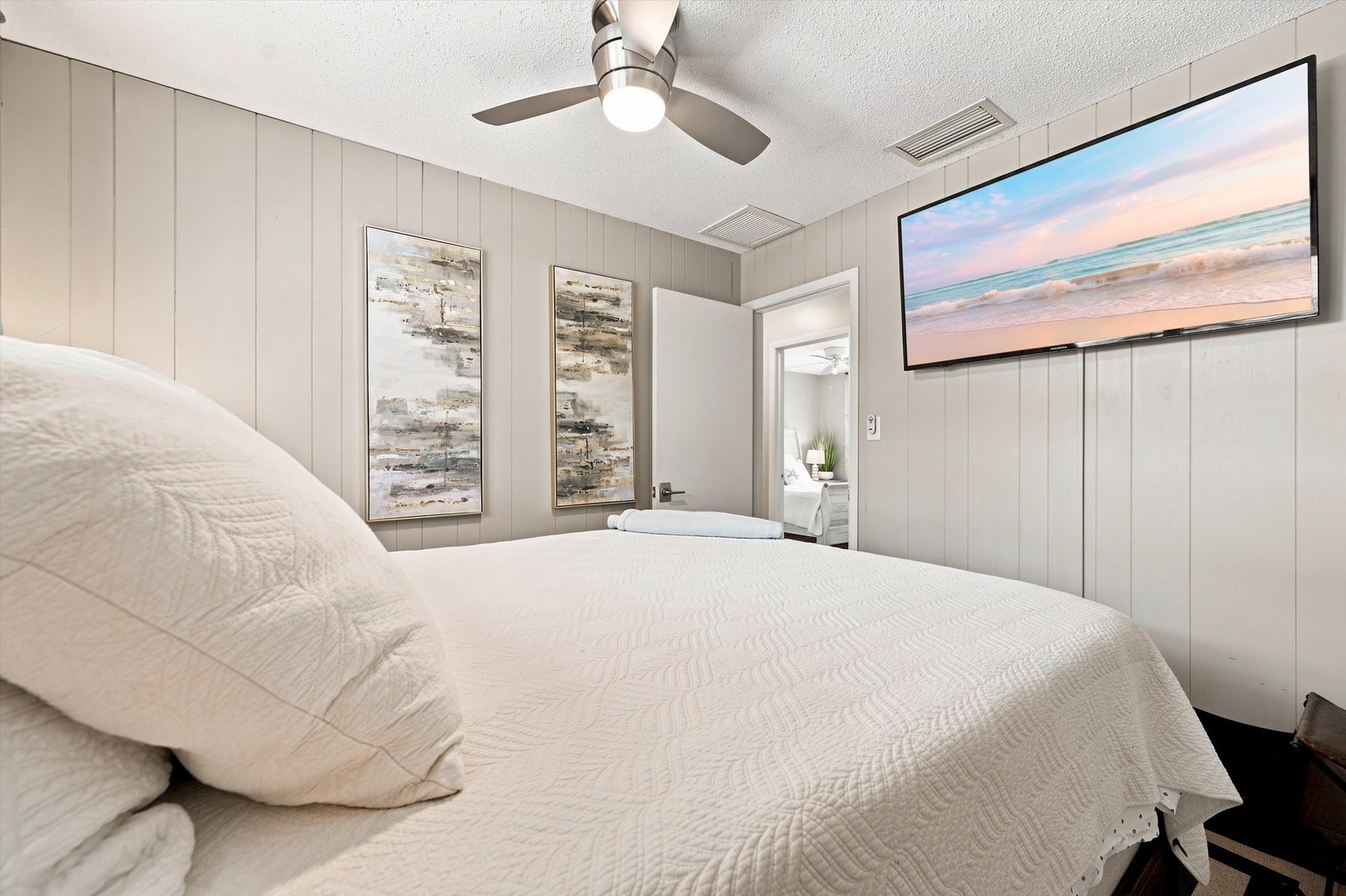 Sink into fresh linens in your serene bedroom, where soft white tones and coastal accents invite restful sleep. The ceiling fan keeps you comfortable year-round, and a glimpse through the doorway reveals a bright en-suite bath.