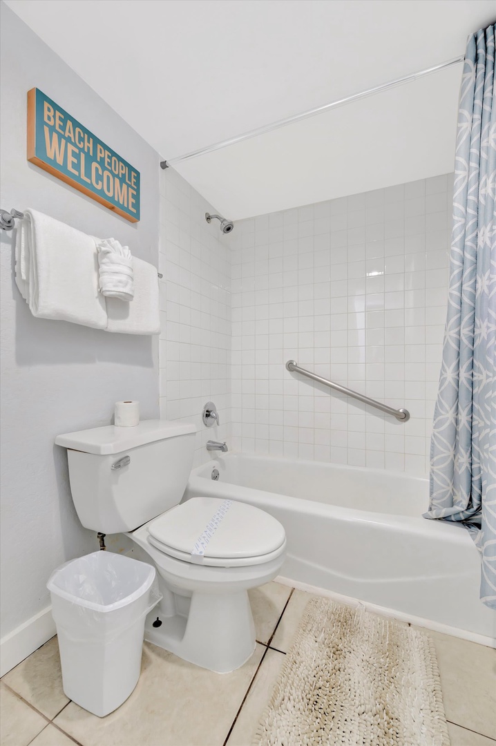 Your bright, tiled bathroom features a tub-shower combo with safety rails and a cheerful beach-themed welcome sign setting a relaxed coastal tone.