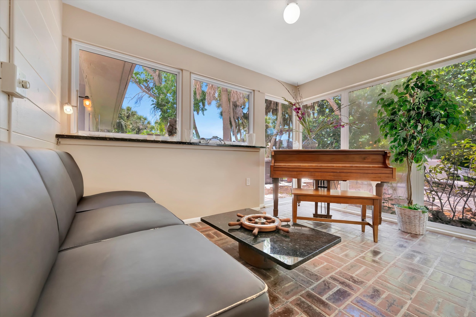 Sun-drenched sunroom with tropical palms framing the windows and grand piano. Brick floors and lush greenery create a peaceful retreat perfect for reading, music, or relaxing.