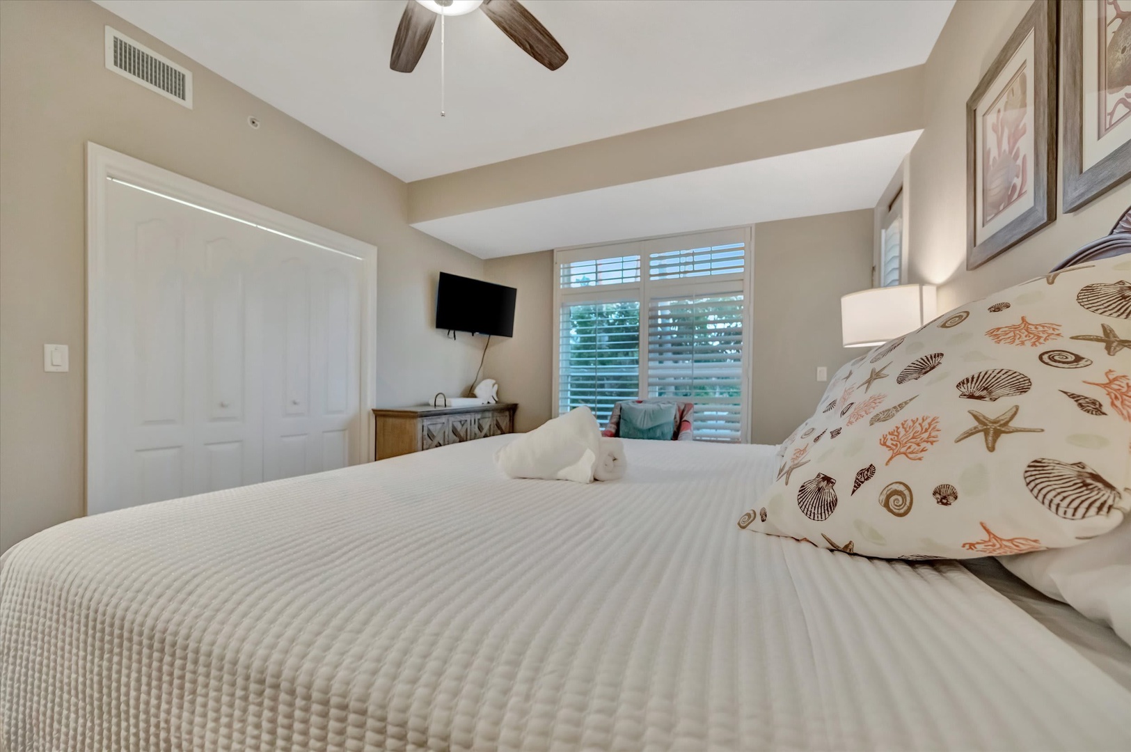 Sink into a plush bed dressed in crisp linens and coastal-themed pillows. Gentle light filters through plantation shutters while a ceiling fan keeps you comfortable. Your peaceful retreat awaits.