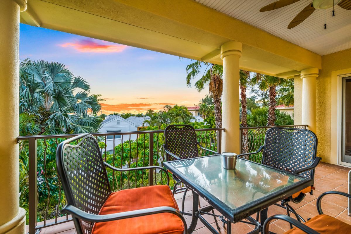 The property's covered balcony features a dining table for four and panoramic views of tropical palm trees and the surrounding neighborhood, with stunning sunsets framing the lush greenery beyond.
