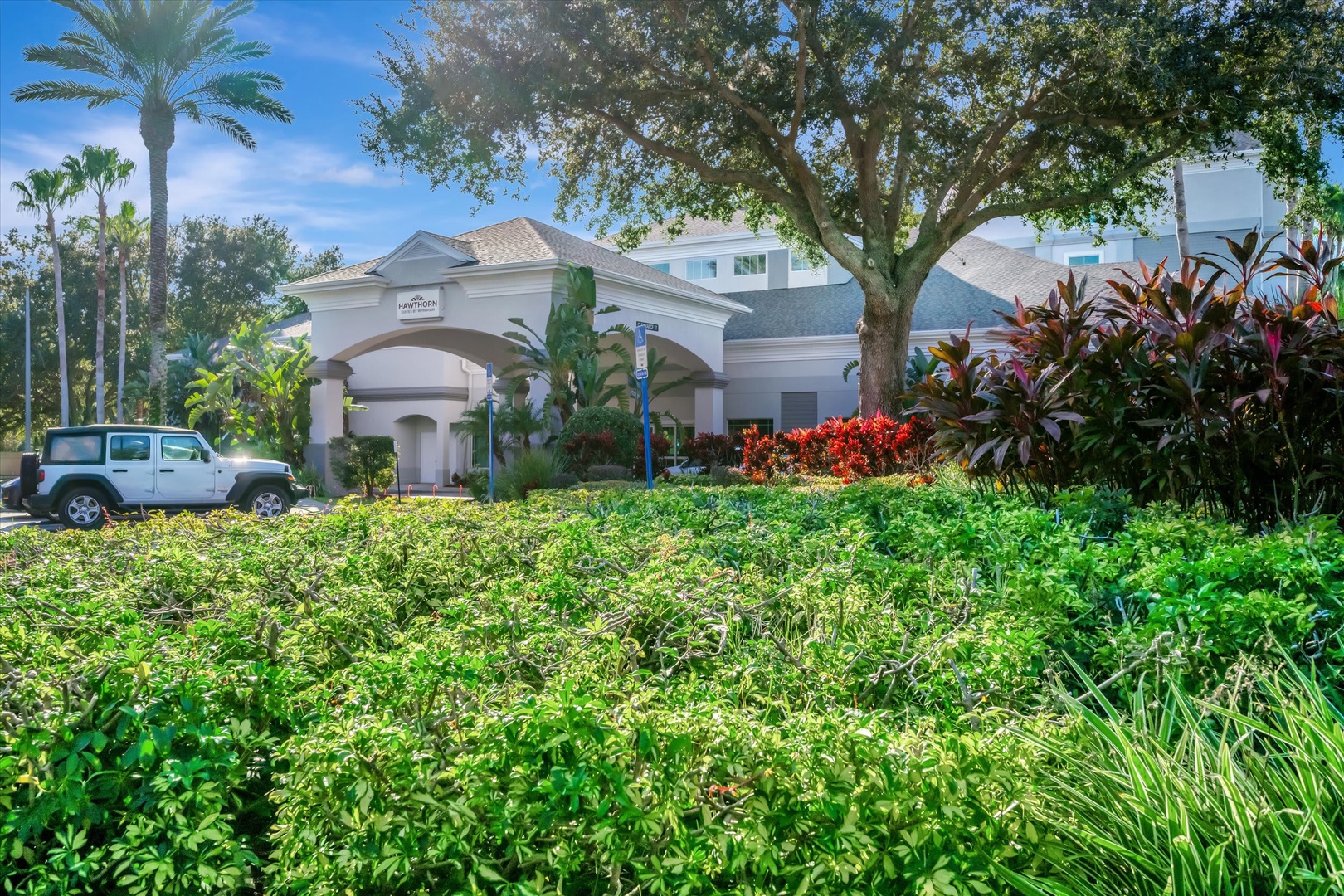 The property features a welcoming entrance framed by vibrant tropical landscaping, with lush palms and colorful plants creating a serene arrival experience under a bright blue sky.