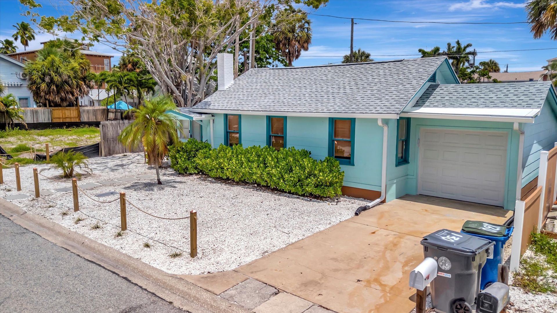 A charming beach cottage with distinctive turquoise exterior, nestled among palm trees in a coastal neighborhood with convenient parking and white gravel landscaping.
