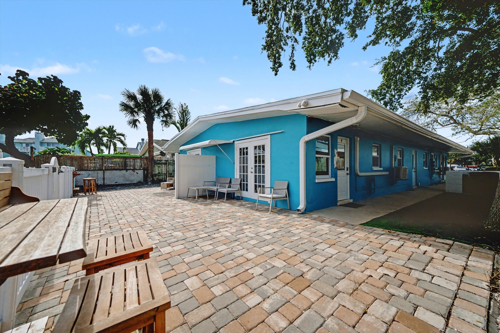 A vibrant turquoise cottage with a spacious brick patio surrounded by palm trees and lush greenery, offering a relaxing outdoor retreat in a sunny tropical setting.