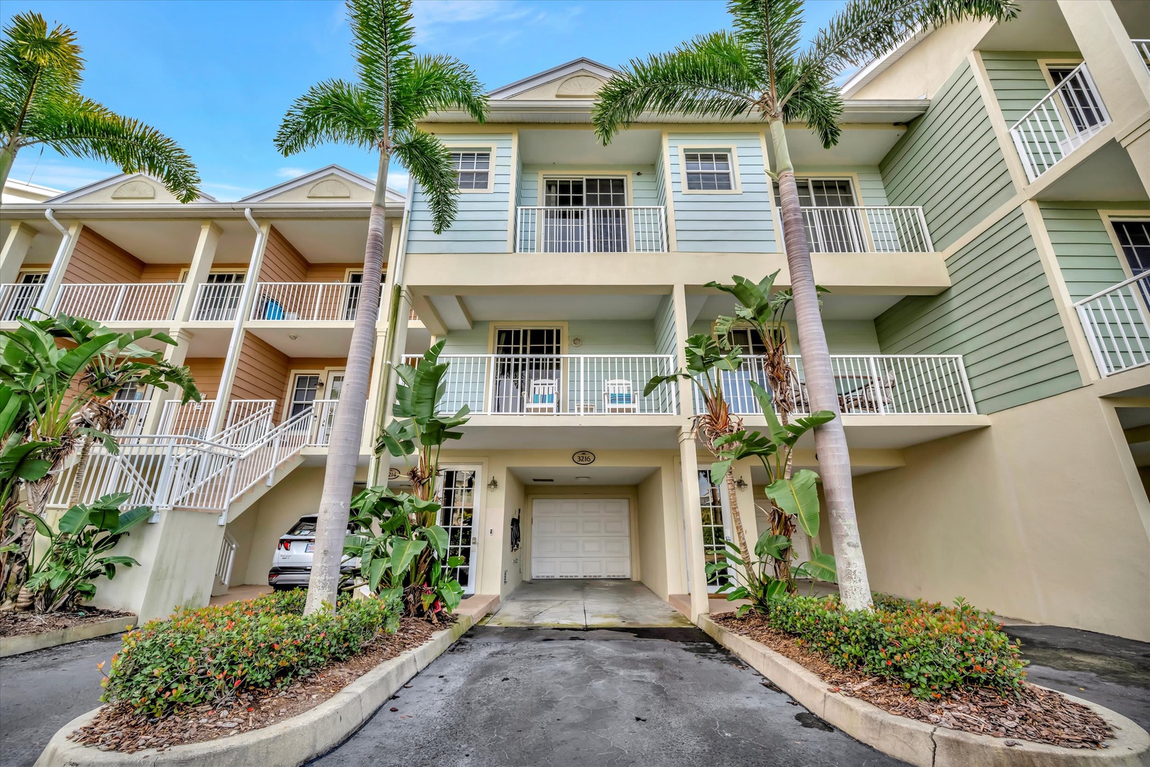 The property features a coastal-style building with spacious private balconies, framed by swaying palms and lush tropical landscaping under sunny skies.